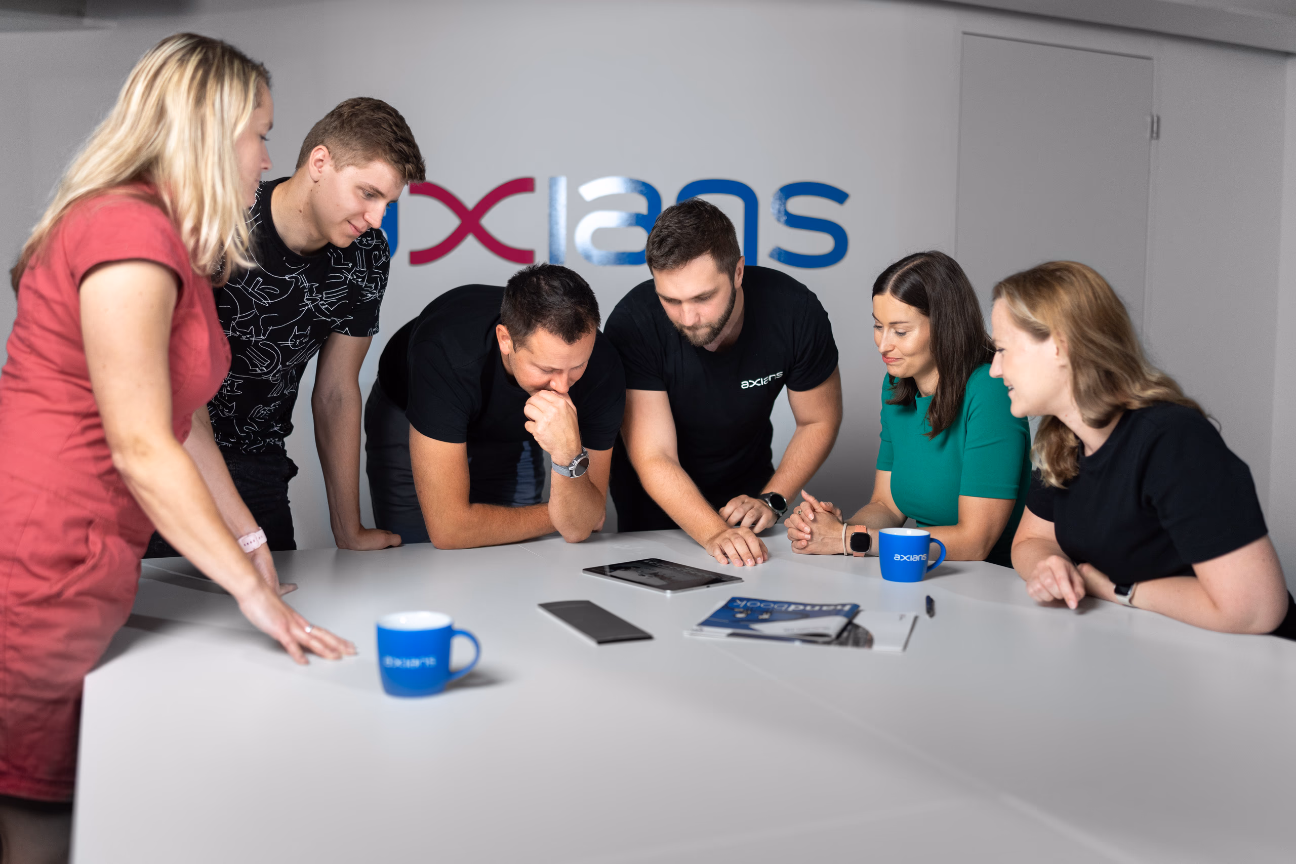 Šest kolegů z Axians nad tabletem u bílého stolu, v pozadí logo firmy na zdi